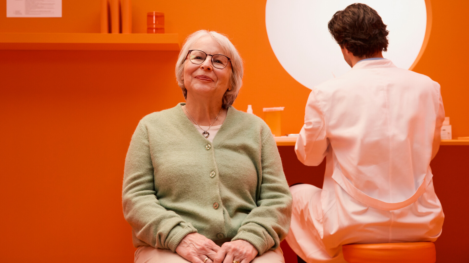 Ältere Patientin sitzt in stilisiertem Impfzimmer. Im Hintergrund bereitet Apotheker Impfung vor. 