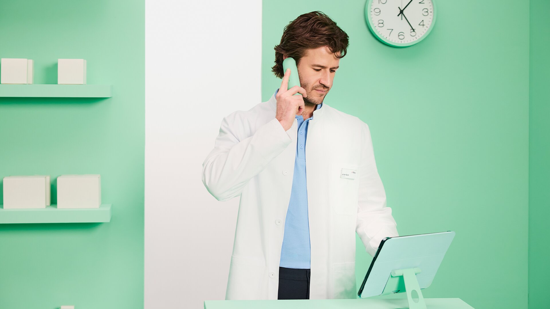 Telefonierender Apotheker steht an Schreibtisch und prüft Informationen am Tablet in stilisierter Apotheke.
