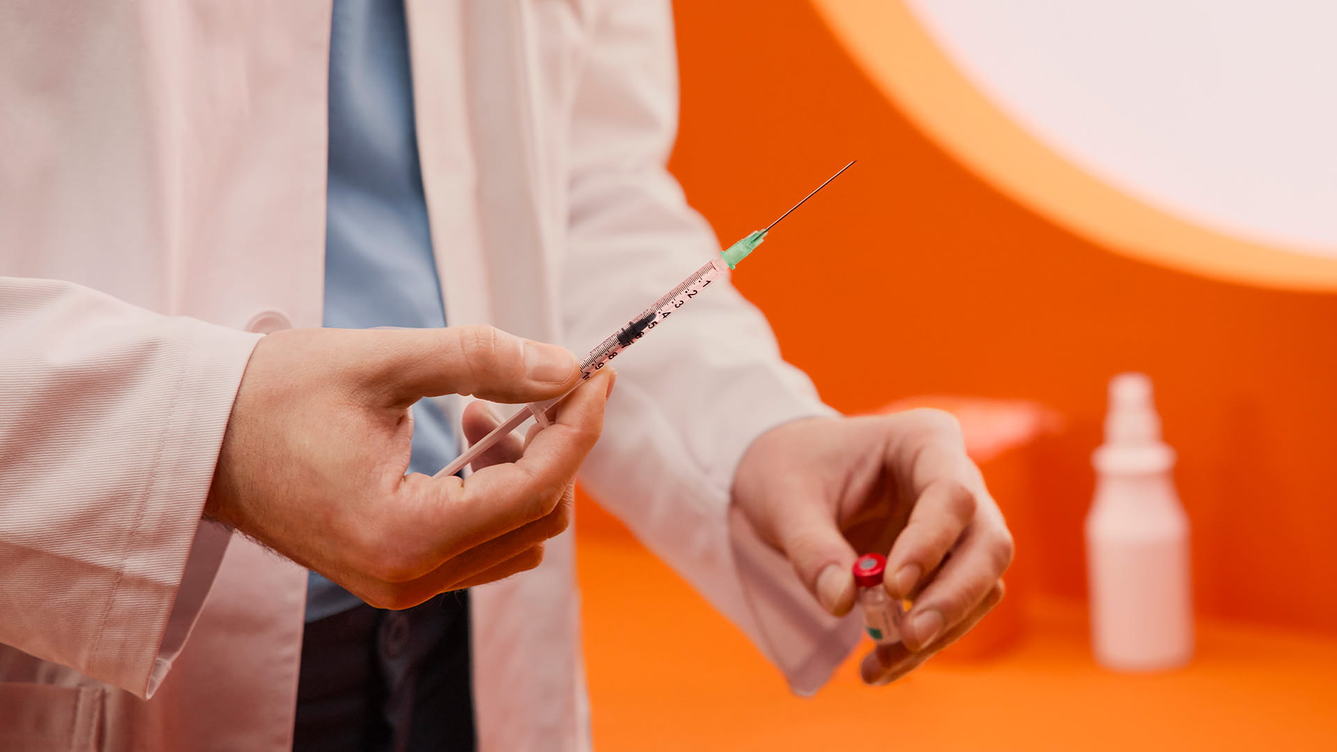 Hände eines Apothekers halten Spritze und Impfstoff-Vial in einem stilisiertem Impfzimmer hoch.