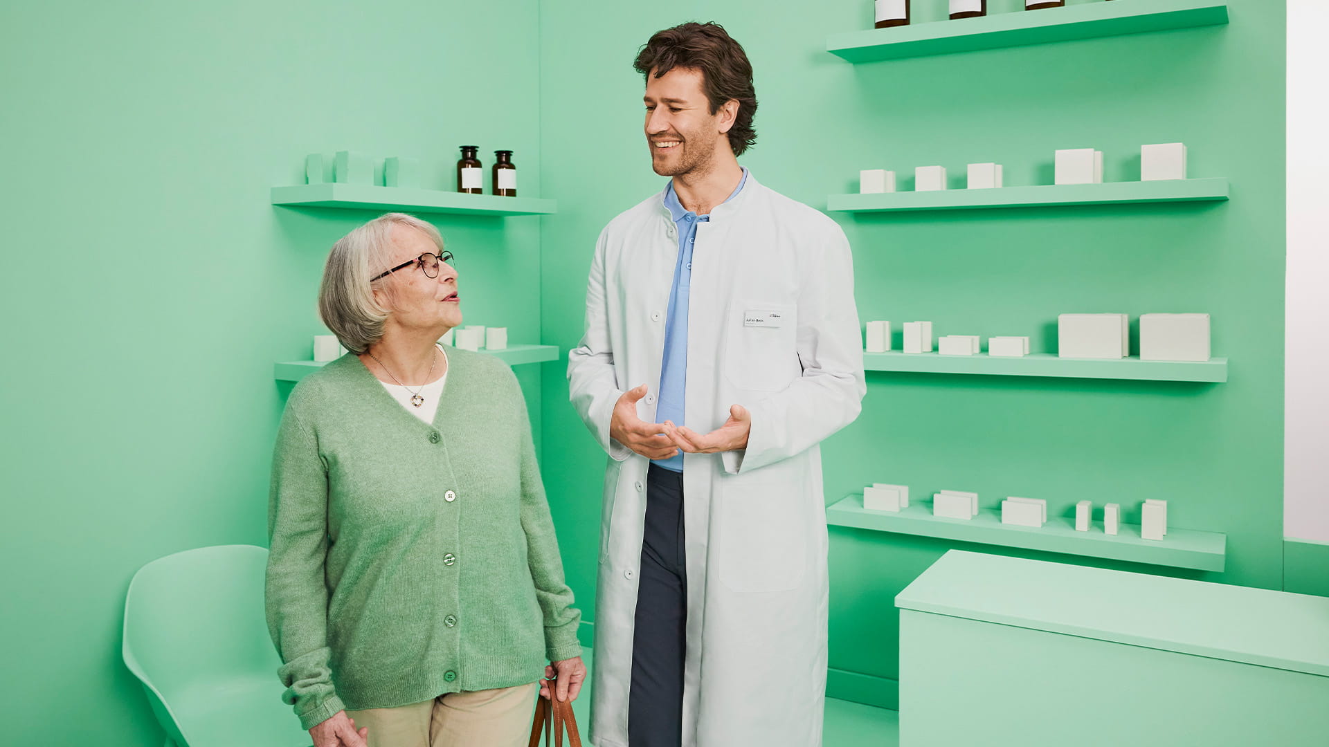 Apotheker und ältere Patientin unterhalten sich stehend in stilisierter Apotheke. 